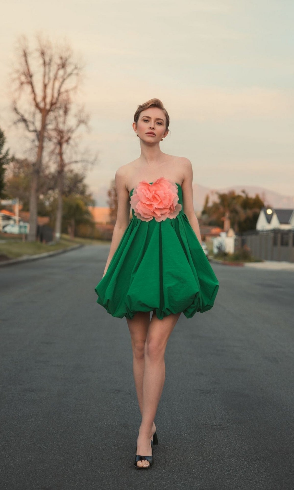 A woman stands on an empty street wearing the Mac Duggal 20931 Short Bubble Party Dress with a large pink flower detail and black open-toe heels.